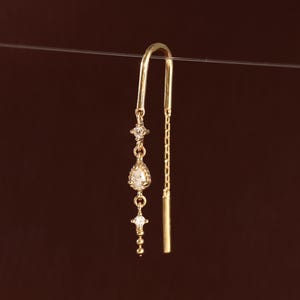 May include: A gold threader earring featuring a delicate chain design with a central pear-shaped stone, two small round accent stones, and a thin metal bar end. The earring is suspended from a thin horizontal wire, showcasing its vertical structure.