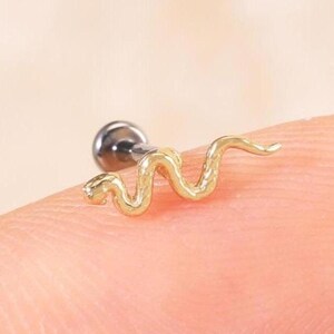 May include: A gold-toned body jewelry stud featuring a small, textured snake-shaped design. The snake is positioned horizontally, with a metallic post and a small circular backing visible behind the decorative front piece, resting on a fingertip.