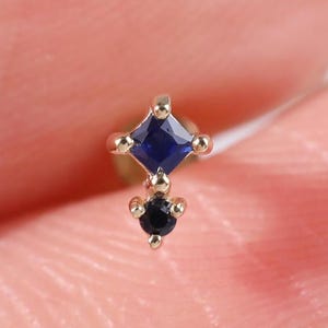 Puede incluir: Un pequeño pendiente dorado con dos piedras preciosas. La piedra superior es una piedra cuadrada de color azul oscuro, y la piedra inferior es una pequeña piedra negra redonda. El pendiente está diseñado con un poste para perforar.