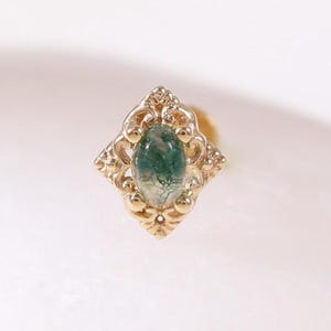 May include: A gold-colored stud earring with an ornate, diamond-shaped setting. The setting surrounds a green gemstone with a moss-like pattern. The earring is set against a white background.