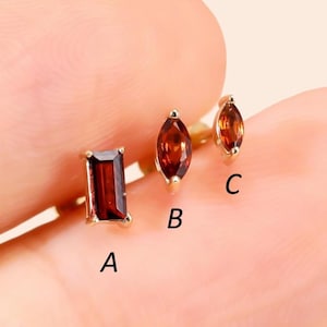 Pode incluir: Três brincos de botão dourados com pedras preciosas castanho-avermelhadas. Os brincos têm três formas diferentes: um retângulo, uma marquesa e uma marquesa menor. Os brincos são exibidos em um fundo claro.