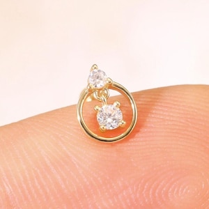 May include: A small, gold-coloured earring featuring two clear, sparkling gemstones. One gemstone is round and set within a small gold circle, the other is teardrop-shaped and positioned above. The earring is shown on a fingertip.