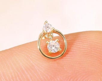 May include: A small, gold-colored earring with two clear, sparkling gemstones. One gemstone is round and set within a small gold circle, while the other is teardrop-shaped and positioned above. The earring is shown on a fingertip.