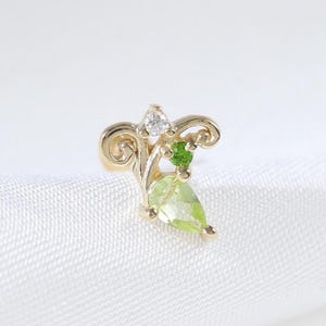 Puede incluir: Un pendiente de color dorado con un diseño de hoja. El pendiente presenta una piedra preciosa verde claro en forma de pera, una pequeña piedra preciosa verde redonda y una piedra preciosa transparente en forma de diamante. El diseño incluye detalles dorados en espiral.