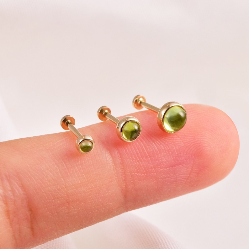 May include: Three gold barbell earrings with green gemstone tops. The earrings are different sizes, with the smallest on the left and the largest on the right.