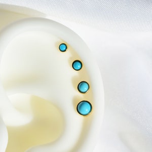 May include: Five turquoise gemstone earrings with gold settings are lined up on a white surface. The earrings are different sizes, with the largest at the bottom and the smallest at the top.