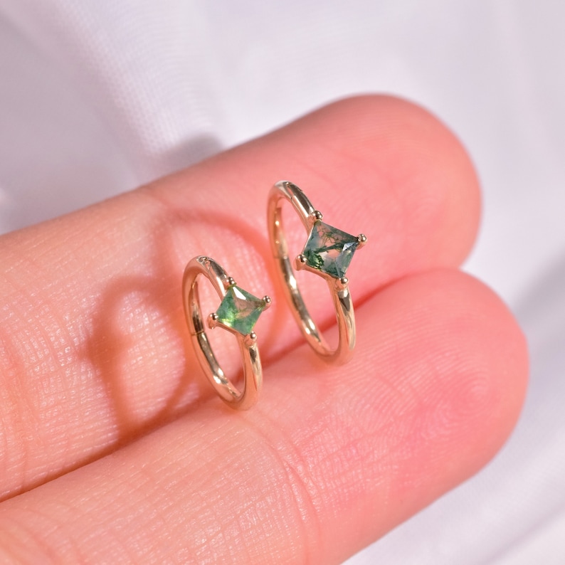 May include: Two gold hoop earrings with green gemstone accents. The earrings are shown on a finger.