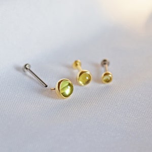 K&ouml;nnte beinhalten: Drei Ohrstecker mit Gold- oder Silberfinish, jeweils mit einem runden, hellgr&uuml;nen Edelstein. Die Ohrringe werden auf einem wei&szlig;en, strukturierten Hintergrund gezeigt. Verschiedene Gr&ouml;&szlig;en.