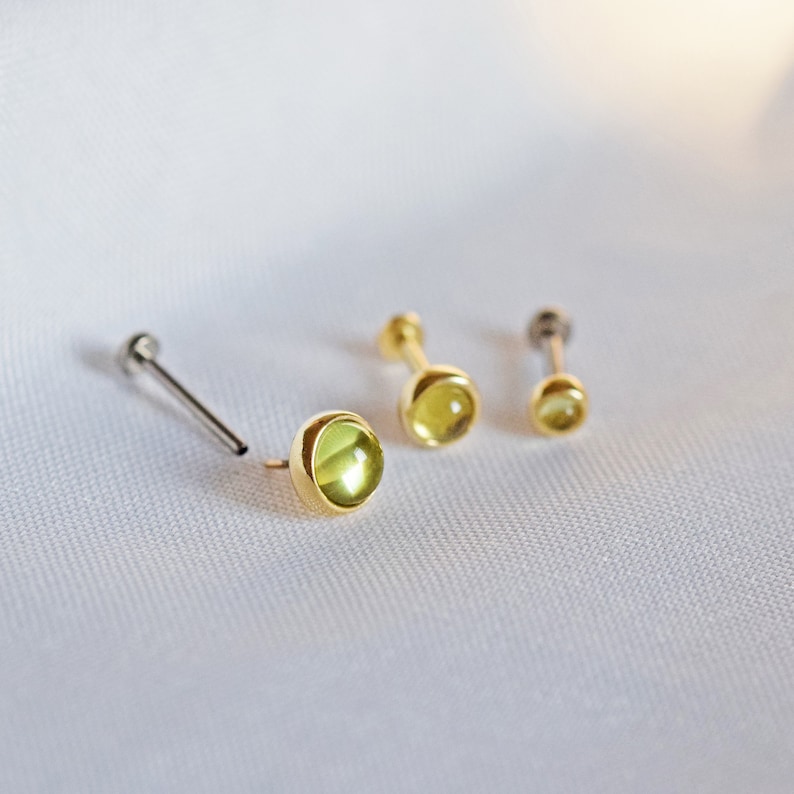 K&ouml;nnte beinhalten: Drei Ohrstecker mit Gold- oder Silberfinish, jeweils mit einem runden, hellgr&uuml;nen Edelstein. Die Ohrringe werden auf einem wei&szlig;en, strukturierten Hintergrund gezeigt. Verschiedene Gr&ouml;&szlig;en.