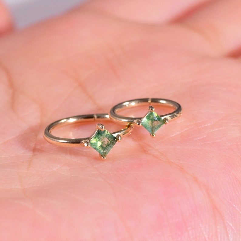 May include: Two gold hoop earrings with small square green gemstones. The earrings are on a hand.