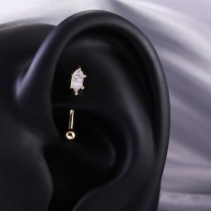 May include: A gold body piercing with a white opal-like stone. The piercing is in a fake ear.