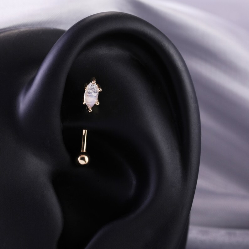 May include: A gold body piercing with a white opal-like stone. The piercing is in a fake ear.