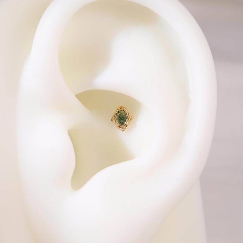 May include: A gold-coloured earring featuring an oval, green gemstone. The earring has an ornate filigree design surrounding the gemstone. The jewellery is displayed on a white ear model, showcasing the intricate details and design.