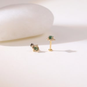 May include: Two gold earrings with green gemstones. One is a flat-backed stud with a square gemstone, and the other is a post earring with a gemstone and a decorative halo. The earrings are displayed on a white surface.