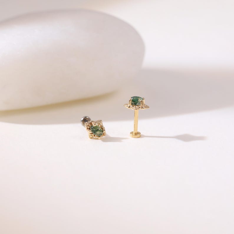 May include: Two gold earrings with green gemstones. One is a flat-backed stud with a square gemstone, and the other is a post earring with a gemstone and a decorative halo. The earrings are displayed on a white surface.