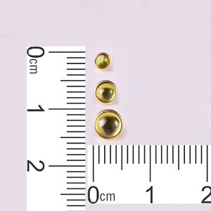 K&ouml;nnte beinhalten: Drei runde, olivgr&uuml;ne Edelsteine in Goldfassungen, vertikal angeordnet. Die Edelsteine variieren in der Gr&ouml;&szlig;e, der gr&ouml;&szlig;te hat einen Durchmesser von etwa 2 cm. Ein Lineal ist zur Ma&szlig;stabsbestimmung neben den Edelsteinen platziert.