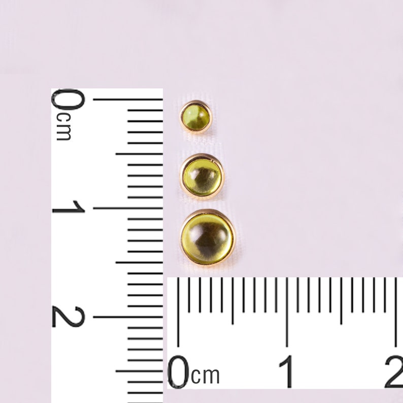 K&ouml;nnte beinhalten: Drei runde, olivgr&uuml;ne Edelsteine in Goldfassungen, vertikal angeordnet. Die Edelsteine variieren in der Gr&ouml;&szlig;e, der gr&ouml;&szlig;te hat einen Durchmesser von etwa 2 cm. Ein Lineal ist zur Ma&szlig;stabsbestimmung neben den Edelsteinen platziert.