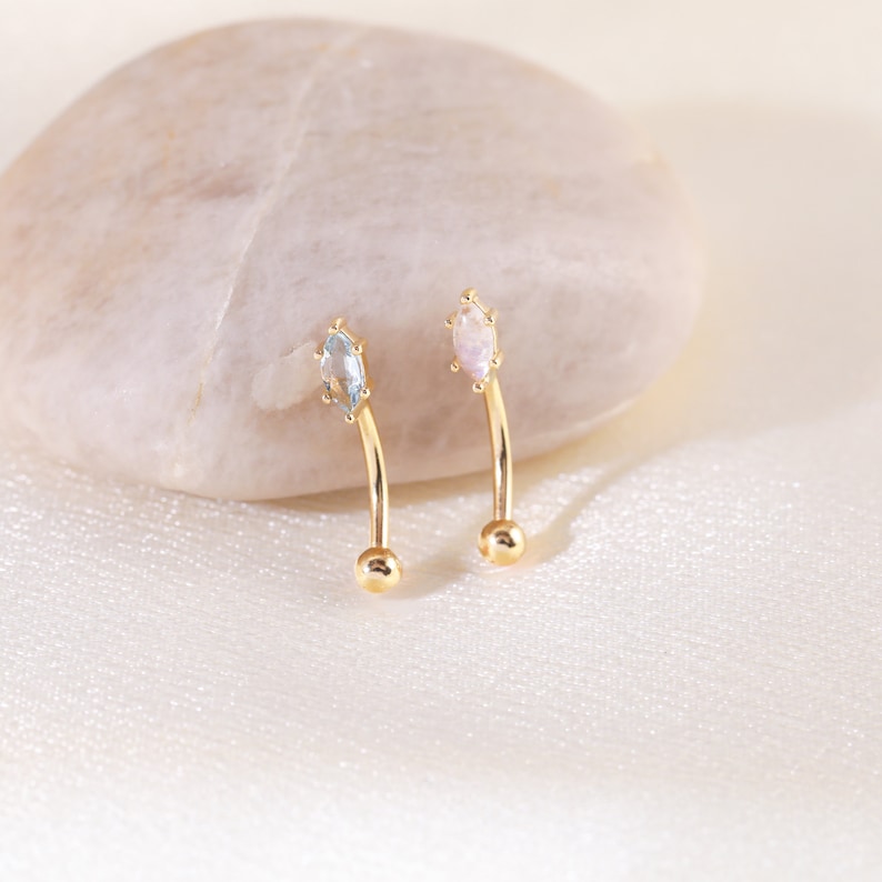 May include: Two gold curved barbell earrings with a marquise shaped gemstone. One earring has a light blue gemstone and the other has a light pink gemstone.