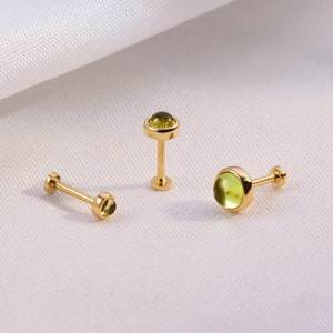 K&ouml;nnte beinhalten: Drei goldfarbene Ohrstecker mit einem kleinen, runden, hellgr&uuml;nen Edelstein. Die Ohrringe haben einen Stecker und einen flachen R&uuml;cken. Die Edelsteine sind in einer L&uuml;nettenfassung gefasst. Die Ohrringe werden auf einer wei&szlig;en, strukturierten Oberfl&auml;che pr&auml;sentiert.