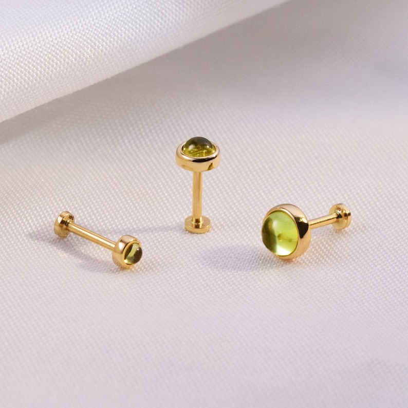 K&ouml;nnte beinhalten: Drei goldfarbene Ohrstecker mit einem kleinen, runden, hellgr&uuml;nen Edelstein. Die Ohrringe haben einen Stecker und einen flachen R&uuml;cken. Die Edelsteine sind in einer L&uuml;nettenfassung gefasst. Die Ohrringe werden auf einer wei&szlig;en, strukturierten Oberfl&auml;che pr&auml;sentiert.