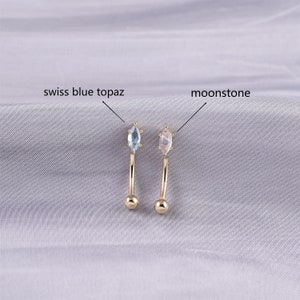 May include: Two gold belly button rings with different gemstones. The ring on the left has a light blue swiss blue topaz gemstone. The ring on the right has a white moonstone gemstone.