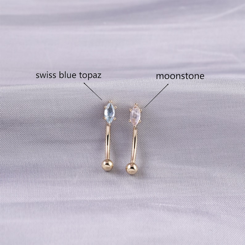 May include: Two gold belly button rings with different gemstones. The ring on the left has a light blue swiss blue topaz gemstone. The ring on the right has a white moonstone gemstone.