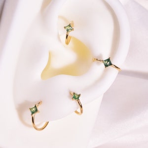 May include: Three gold hoop earrings with green square-shaped gemstones. The earrings are shown on a white background.