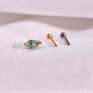 K&ouml;nnte beinhalten: Drei Schmuckst&uuml;cke werden auf einer wei&szlig;en Oberfl&auml;che pr&auml;sentiert. Eines ist ein goldener Ohrstecker mit einem gr&uuml;nen, tropfenf&ouml;rmigen Stein. Zwei weitere gold- und silberfarbene Ohrstecker sind ebenfalls abgebildet. Der Schmuck ist wahrscheinlich f&uuml;r Ohrpiercings.