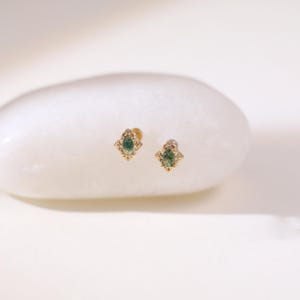 May include: A pair of gold stud earrings featuring a green gemstone. The earrings have an ornate design around the central stone. The earrings are presented on a white, rounded surface.