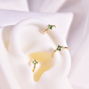 May include: Three gold hoop earrings with small square green gemstones. The earrings are displayed on a white surface.