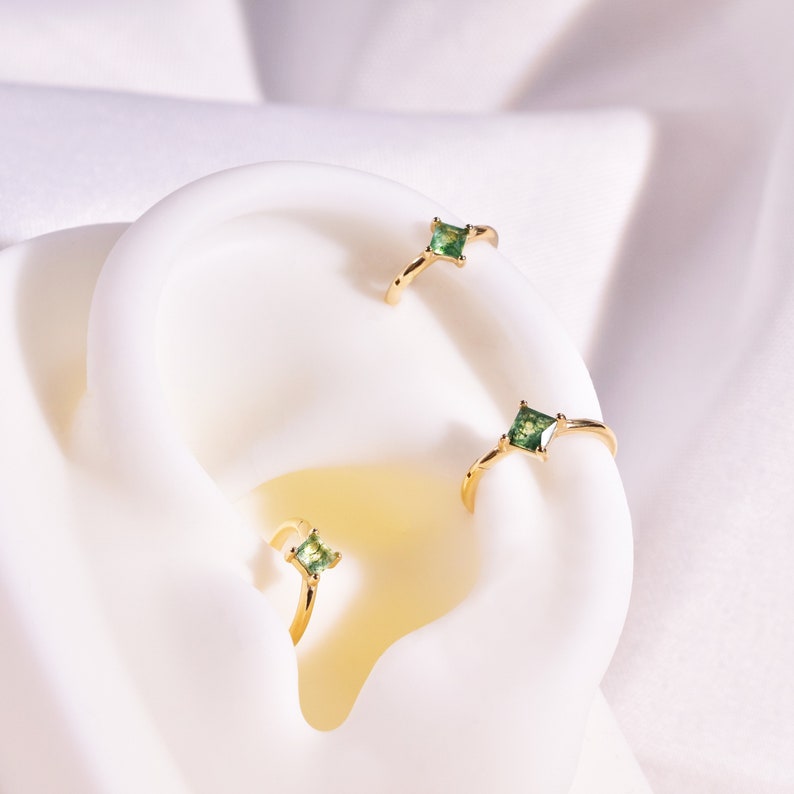 May include: Three gold hoop earrings with small square green gemstones. The earrings are displayed on a white surface.