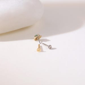 May include: A selection of small earrings and posts. One earring has a gold setting with a small, green gemstone. Two posts are visible, one gold and one silver, offering interchangeable earring options.