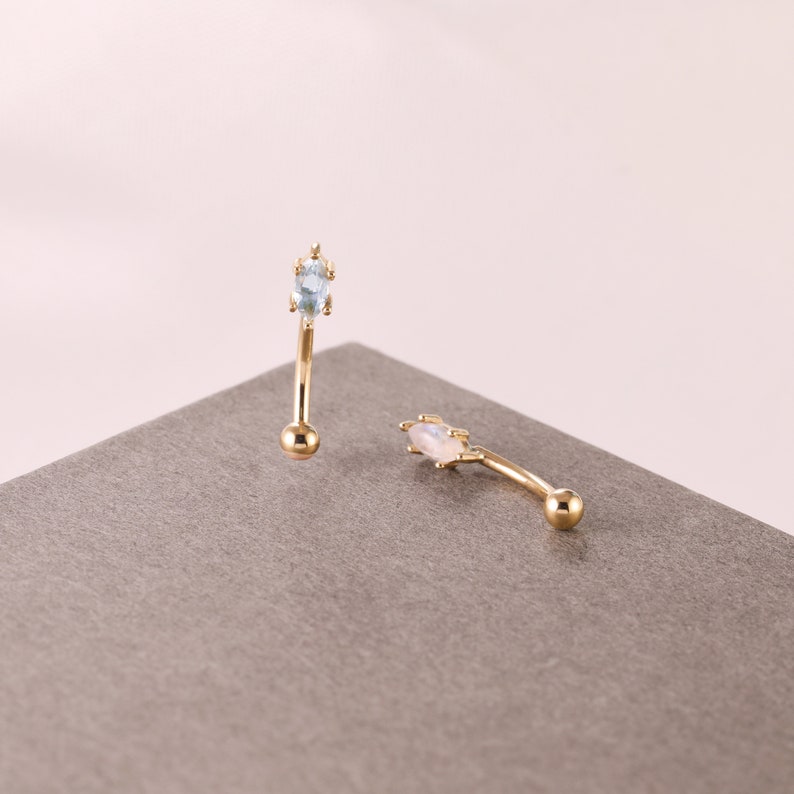May include: Two gold body jewelry pieces with a curved barbell design. One piece features a light blue gemstone and the other features a white gemstone.