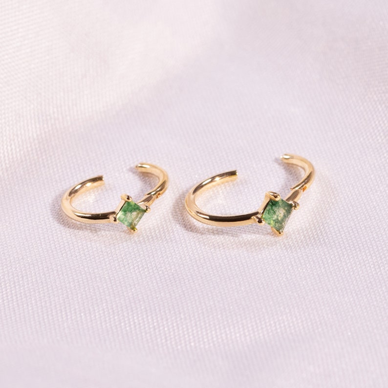 May include: A pair of gold ear cuffs with small square green gemstones.