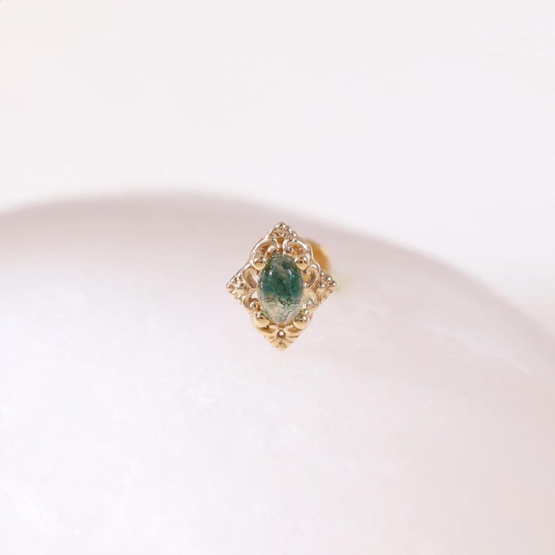 May include: A gold-colored stud earring with an ornate, diamond-shaped setting. The setting surrounds a green gemstone with a moss-like pattern. The earring is set against a white background.