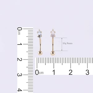 May include: Two gold-colored curved barbell body jewelry pieces with clear gemstones. The jewelry is 8mm in length and 16 gauge. A ruler is in the background for size reference.