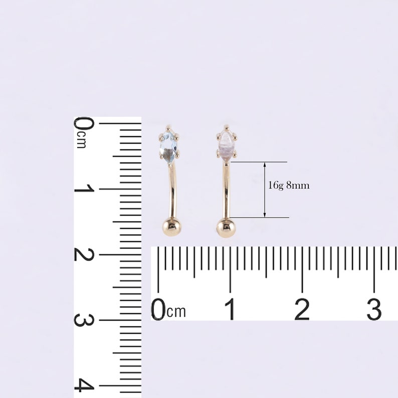 May include: Two gold-colored curved barbell body jewelry pieces with clear gemstones. The jewelry is 8mm in length and 16 gauge. A ruler is in the background for size reference.
