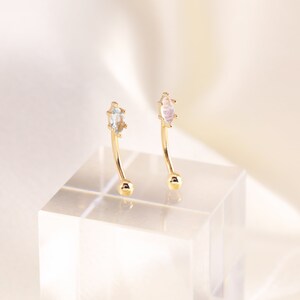 May include: Two gold curved barbell earrings with a clear gemstone on one and a white iridescent gemstone on the other.
