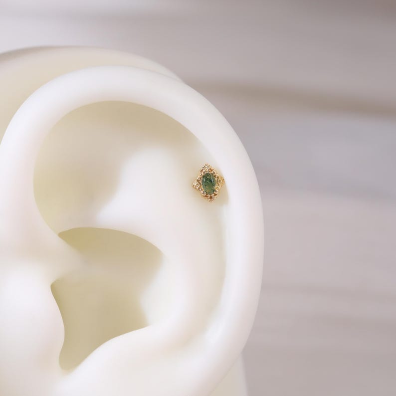 May include: A gold-coloured earring featuring a green gemstone set within a decorative, diamond-shaped frame. The earring is displayed on a white ear model, highlighting its design. The gemstone is slightly oval in shape.