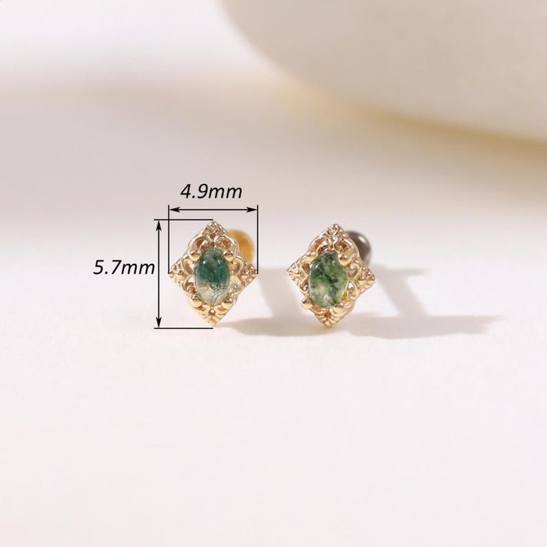 May include: A pair of gold-coloured stud earrings. Each earring features a green gemstone in a decorative, diamond-shaped setting. The earrings measure 5.7mm in height and 4.9mm in width. The gemstones have a teardrop shape.