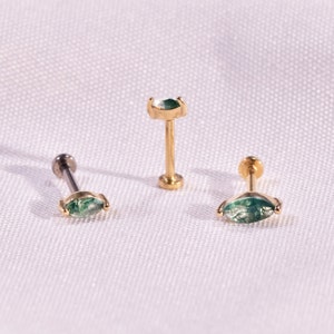 K&ouml;nnte beinhalten: Drei Schmuckst&uuml;cke: zwei goldfarbene Ohrringe mit einem gr&uuml;nen, marquisef&ouml;rmigen Stein und ein goldfarbener Ohrstecker mit einem gr&uuml;nen, marquisef&ouml;rmigen Stein. Die Ohrringe werden auf einer wei&szlig;en Oberfl&auml;che pr&auml;sentiert.