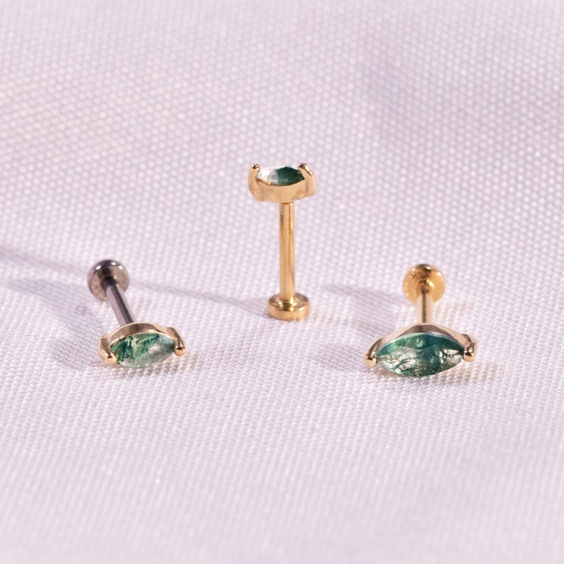 K&ouml;nnte beinhalten: Drei Schmuckst&uuml;cke: zwei goldfarbene Ohrringe mit einem gr&uuml;nen, marquisef&ouml;rmigen Stein und ein goldfarbener Ohrstecker mit einem gr&uuml;nen, marquisef&ouml;rmigen Stein. Die Ohrringe werden auf einer wei&szlig;en Oberfl&auml;che pr&auml;sentiert.