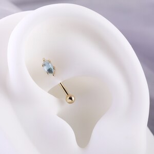 May include: A gold barbell with a light blue marquise-shaped gemstone. The barbell is shown in a close-up view on a white mannequin ear.