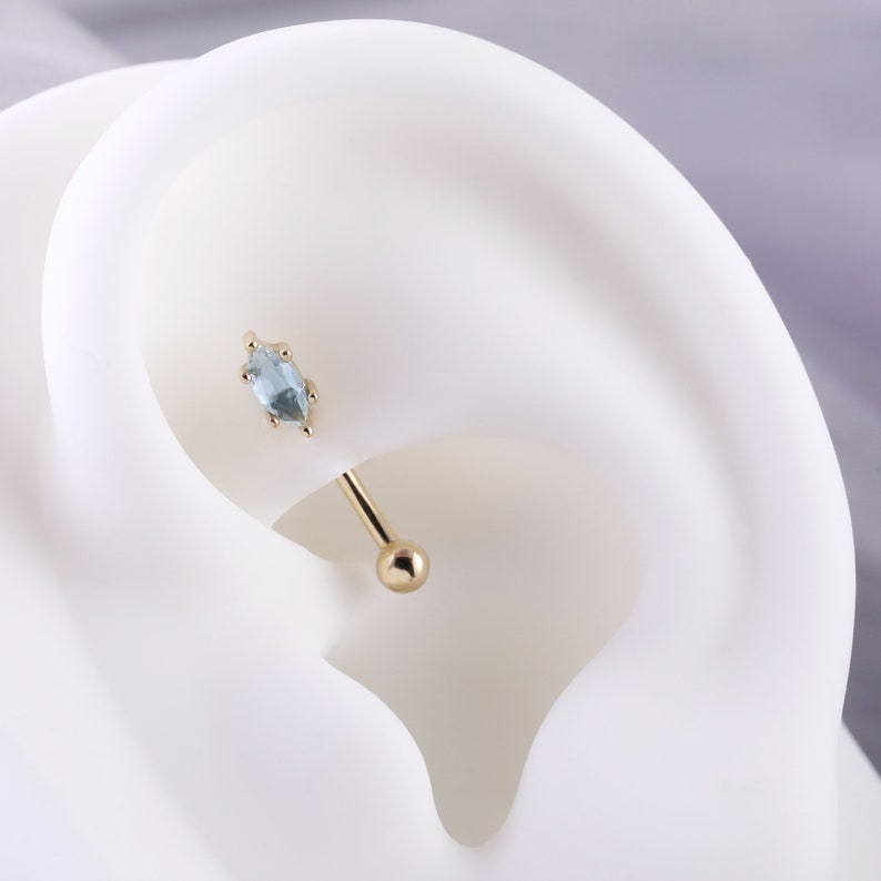 May include: A gold barbell with a light blue marquise-shaped gemstone. The barbell is shown in a close-up view on a white mannequin ear.