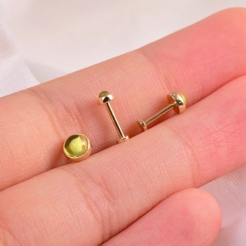 K&ouml;nnte beinhalten: Drei goldfarbene Ohrstecker mit einem hellgr&uuml;nen Edelstein. Zwei Ohrringe sind im Stecker-Stil, einer ist ein einzelner Edelstein. Die Ohrringe werden auf einer hellen Oberfl&auml;che pr&auml;sentiert.