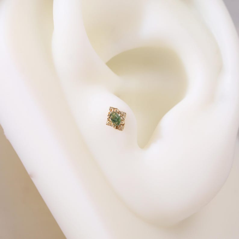 May include: A gold-coloured square earring with a central green gemstone. The earring is set with smaller, clear stones around the central gem. The jewellery is displayed on a white ear model, highlighting the design and details of the earring.