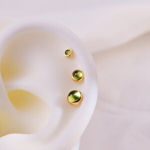 K&ouml;nnte beinhalten: Drei goldene Ohrstecker mit runden, hellgr&uuml;nen Edelsteinen werden auf einer wei&szlig;en Oberfl&auml;che gezeigt. Die Ohrringe sind unterschiedlich gro&szlig;, der gr&ouml;&szlig;te unten und der kleinste oben. Die Edelsteine haben eine subtile, durchscheinende Qualit&auml;t.