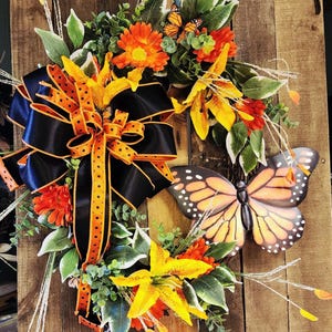 May include: A decorative wreath featuring a large black bow with orange polka dot ribbon, surrounded by orange and yellow flowers, green leaves, and a large butterfly. The wreath is mounted on a wooden background.
