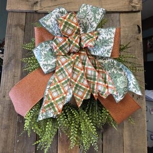 May include: A decorative bow with a mix of patterned ribbons and greenery. The bow features a large, glittery orange ribbon, floral print ribbons, and plaid ribbons in green, orange, and white. The bow is set against a wooden background.