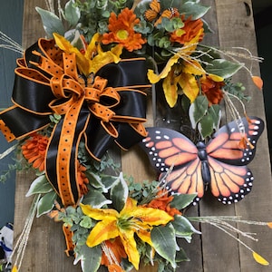 Könnte beinhalten: Ein dekorativer Kranz mit orangefarbenen und gelben Blumen, grünen Blättern und einer großen schwarzen Schleife mit orangefarbenen Punkten. Ein großer Schmetterling und kleinere Schmetterlingsakzente vervollständigen das Design. Der Kranz wird auf einem Holzgrund gezeigt.